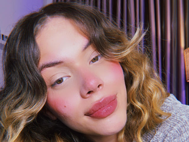 Close-up photo of EmmaRiveraa, light-skinned with wavy brown and blonde hair, full pink lips slightly parted, soft expression, dark eyebrows, slight blush on cheeks, dimly lit background with vertical lines.
