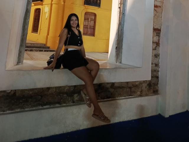 TalianaSquirt, sexy con un atuendo negro, sentada en un borde blanco, con las piernas separadas, un pie levantado, un edificio amarillo de fondo, oscuro. sombras, pose seductora, expresión confiada, noche, entorno urbano, informal, provocativo..