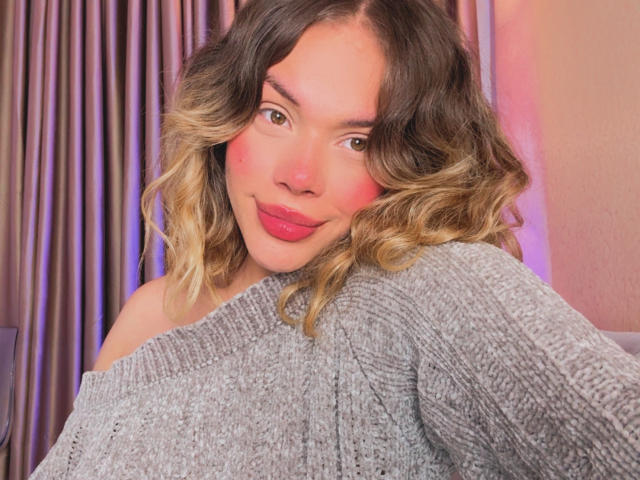 Fotografía de EmmaRiveraa, primer plano, cabello ondulado rubio y castaño, lápiz labial rojo, suéter gris con un hombro descubierto, ligera sonrisa. Iluminación rosa y azul en el fondo, tensión sexual, insinuación de pene en la pose..