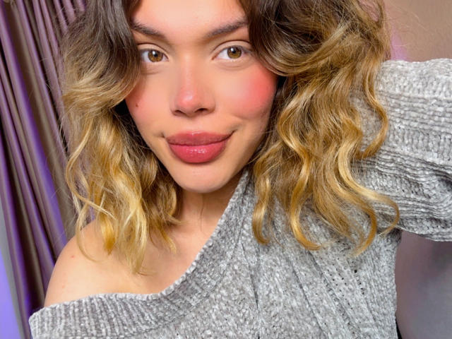 Close-up photo of EmmaRiveraa, brown-eyed woman with wavy blonde hair, wearing an off-shoulder gray sweater. She has a slightly flushed face, red lips, and a teasing smile. Her hand rests on her head.