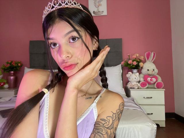 Fotografía de KeityRousse, una mujer asiática con cabello largo y negro trenzado, que lleva una blusa blanca de encaje y una tiara de plata. mano en la mejilla, tatuaje en el brazo superior, dormitorio rosa.