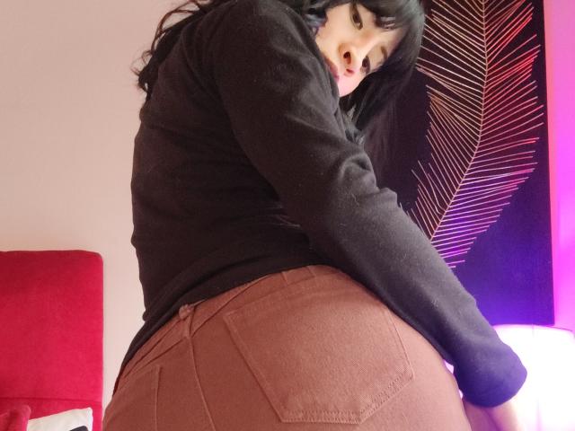 Fotografía de IssisDiosa con cabello largo y negro, vistiendo un suéter negro y pantalones marrones ajustados, inclinándose hacia adelante. Mirada seductora, que resaltaba sus redondeados y prominentes glúteos. Fondo rojo brillante y morado..