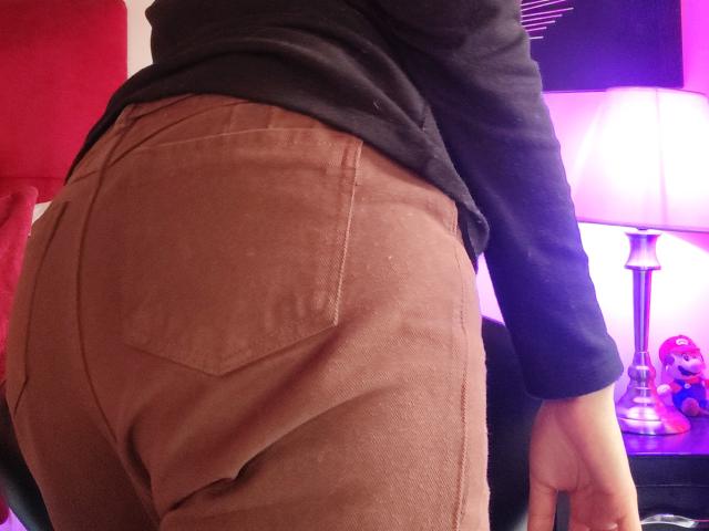 Fotografía de primer plano del trasero de IssisDiosa, que lleva pantalones de pana marrones con bolsillos visibles. Camiseta negra, mano apoyada sobre una superficie negra. Fondo: luces brillantes de color púrpura y blanco, tela roja. Pose sugestiva, sensual..