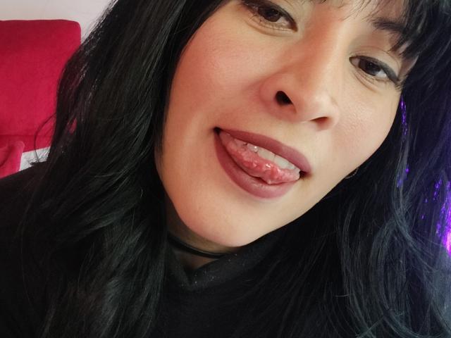 Fotografía de primer plano de IssisDiosa, una joven con cabello largo y negro, que saca la lengua con una expresión juguetona. Tiene la piel clara, ojos oscuros y un ligero rubor. Hay un cojín rojo en el fondo..