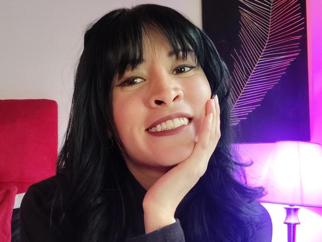 Photograph of IssisDiosa, smiling with hand on cheek, black hair, bangs, brown eyes, wearing black top, red cushion in background, purple and black patterned wall, soft lighting, slight sexual tension.