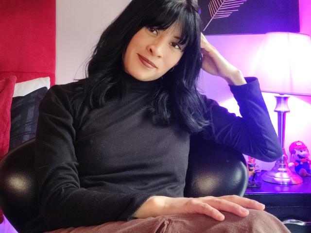 Fotografía de IssisDiosa, una mujer con cabello negro y flequillo, vestida con un suéter negro de cuello alto y pantalones marrones, sentada con con la mano en el pelo, con luces de neón brillantes al fondo. Increíblemente atractivo/a..