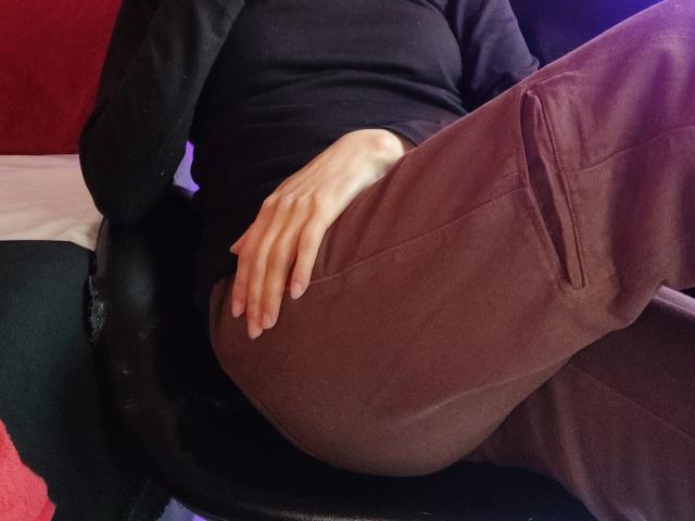 Gros plan d'IssisDiosa assise, portant un haut noir et un pantalon bordeaux. La main repose sur la cuisse, créant un léger renflement dans le pantalon. Cadre intime et sombre avec un fond rouge et noir..