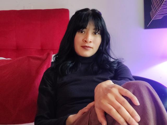 Photographie d'IssisDiosa, assise sur un canapé rouge, portant un pull noir à col roulé et un pantalon violet, avec une main reposant sur son genou, cheveux noirs, regard intense, tension sexuelle.