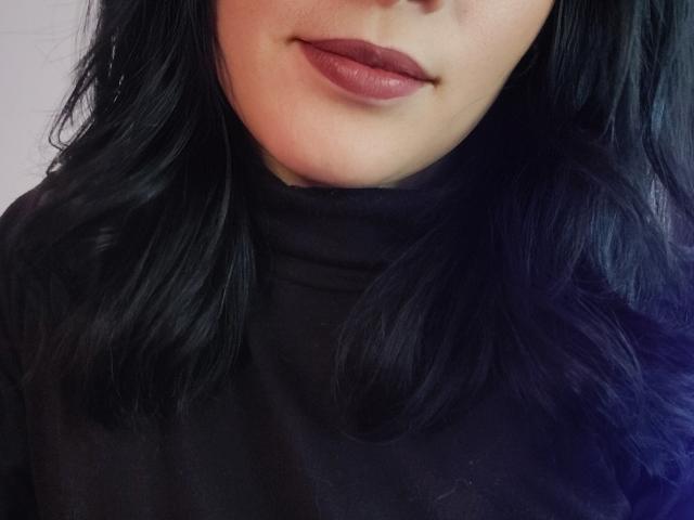 Gros plan du visage d'IssisDiosa, montrant des cheveux foncés et ondulés, des lèvres pulpeuses avec du rouge à lèvres rouge foncé, et un léger sourire. Elle porte un haut noir à col montant, dégageant une aura sexy et sensuelle..