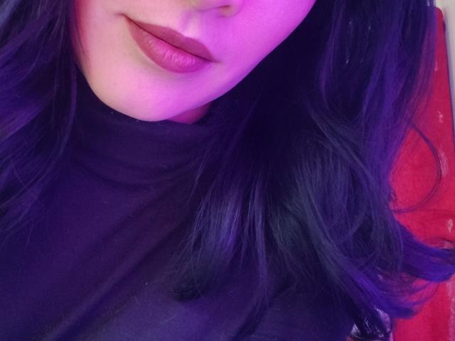 Primo piano del viso e del collo di IssisDiosa, che mostra labbra piene con rossetto rosa-viola, capelli neri parzialmente visibili. Indossava un maglione nero a collo alto. Sfondo rosso. Luci viola e blu che esaltano la pelle. Incredibilmente sexy..