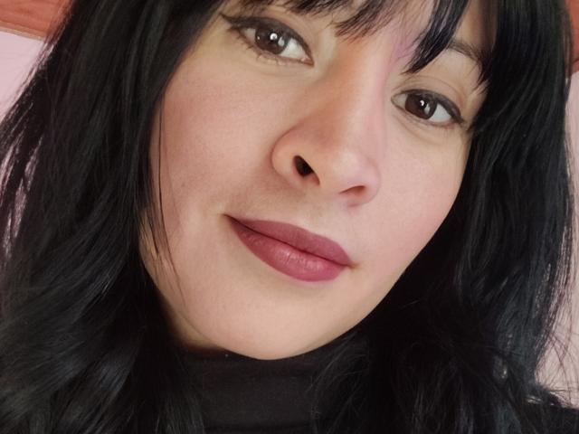 Close-up photo of IssisDiosa, a fair-skinned woman with black hair and bangs, wearing dark red lipstick. She has a subtle smile, brown eyes, and slight makeup. Cock not visible.