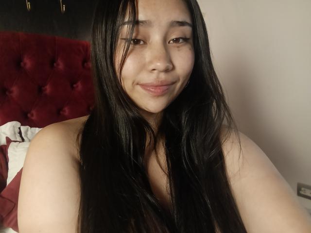 Close-up of Karollteyes with long, straight black hair, smiling slightly, sitting on a bed with a red, tufted headboard. Light skin, casual, slightly messy, sexy vibe, soft lighting.