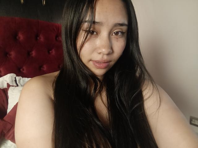 Photograph of Karollteyes, an Asian woman with long black hair, bare shoulders, and a slight smile, sitting in front of a red tufted headboard. She exudes sexual tension with direct eye contact.