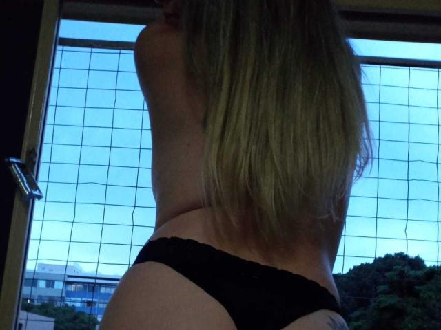 A BadMah está de volta, com cabelo loiro, calcinha de renda preta, tatuagem visível na parte inferior das costas, em pé perto da janela com uma grade. Céu azul lá fora, efeito de silhueta, leve sombra nas costas..