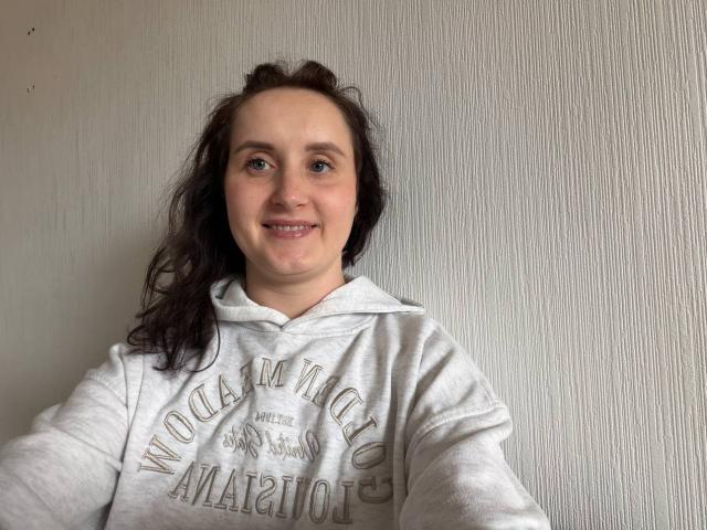 AmeliaStoneX met nat, golvend zwart haar, lachend, gekleed in een witte hoodie met de tekst "All Star", leunt tegen een behandelde, lichtgrijze muur. Zacht licht accentueert haar ontspannen, informele houding..