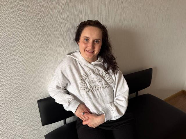 AmeliaStoneX met krullend zwart haar, lachend, draagt een witte hoodie, handen rusten op een zwarte stoel, witte muur met textuur. Op de achtergrond, zwarte broek, subtiele seksuele spanning, nonchalante pose..