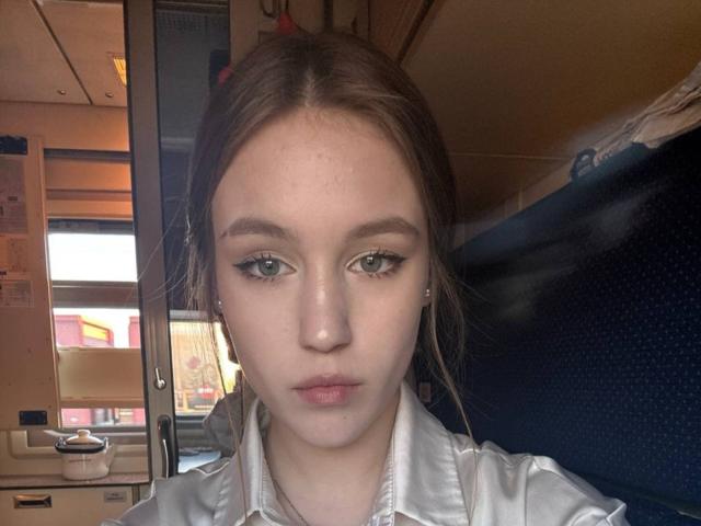 Close-up photograph of CorinaMakinster, a young woman with fair skin, green eyes, and brown hair, wearing a white shirt, looking directly at the camera with a neutral expression. Background includes indoor lighting and shelves.