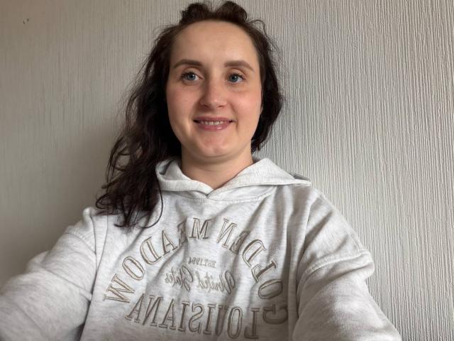 AmeliaStoneX mit zerzaustem, schwarzem Haar, lächelnd, trägt eine weiße Kapuzenjacke mit dem Aufdruck "Rogue Athletics". Weiches Licht, leicht strukturierter weißer Hintergrund. Lässige, entspannte Pose, leichte sexuelle Spannung..