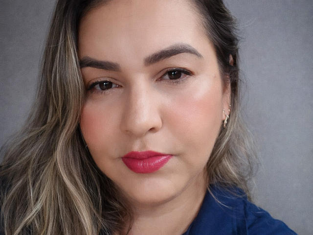 Close-up of BadMah with fair skin, brown eyes, and red lipstick. She has wavy brown hair, dark eyebrows, and wears a dark blue shirt. She's looking directly at the camera with a slight smirk.