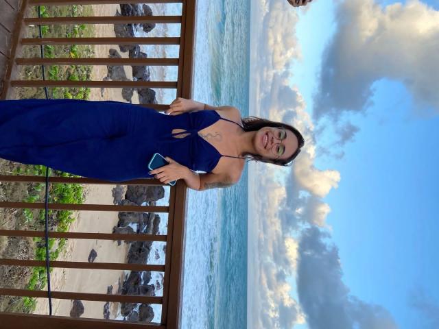 SecretLust-hot staat op een houten veranda, gekleed in een diepblauwe, diep uitgesneden jumpsuit die haar decolleté laat zien. De oceaan en een bewolkte lucht op de achtergrond. Ze houdt een blauwe telefoon vast en glimlacht zelfverzekerd. Heel erg aantrekkelijk..