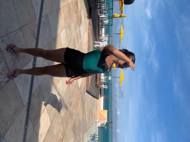 Photographie d'une plage ensoleillée, avec SecretLust-hot en premier plan. Elle porte un débardeur vert et un short noir, les bras levés, projetant des ombres sur le trottoir. Incroyablement sexy..