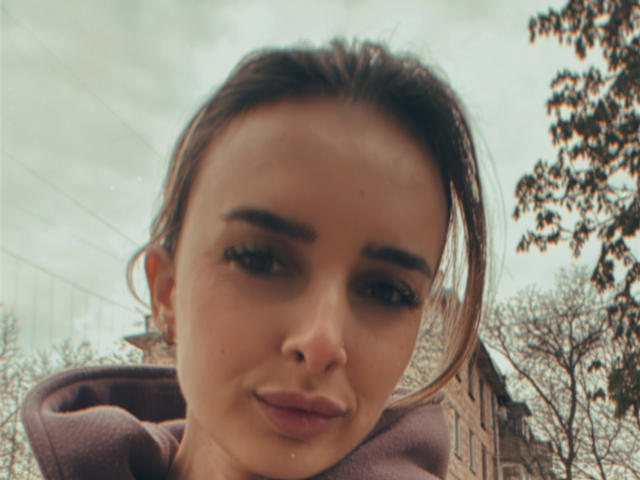 Close-up foto van KatrinaHotX69 met een zachte glimlach, bruin haar naar achteren gebonden, gekleed in een bruine hoodie. De achtergrond bestaat uit bomen en een gebouw. De lucht is licht bewolkt. De seksuele spanning in haar blik..