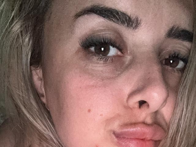 Close-up photo of KatrinaHotX69's face with thick, dark eyebrows, brown eyes, and full lips. Her blonde hair partially covers the right side. She's looking slightly upward with a sultry expression.