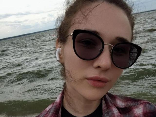 Primer plano de NatoshaBurdine cerca del agua, con gafas de sol negras, auriculares blancos y una camisa roja de cuadros. Su cabello está despeinado, los labios ligeramente fruncidos, y se ven olas del mar al fondo. Mirada muy seria..