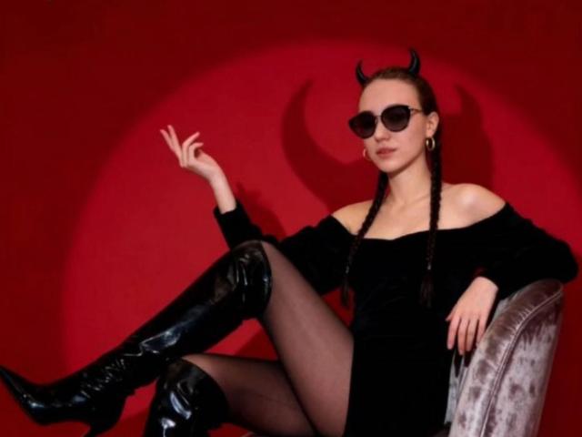 Natosha Burdine sitzt auf einem grauen Stuhl, trägt ein schwarzes Kleid mit freiem Schulterbereich, schwarze hohe Stiefel und eine schwarze Sonnenbrille. mit Teufelshörnern, die rechte Hand erhoben, roter Hintergrund, sexy und selbstbewusst.