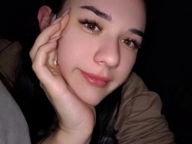 Primer plano de NatoshaBurdine, de piel clara, con cabello oscuro, ojos marrones y labios carnosos, con la mano apoyada en la mejilla. Mirando a la cámara con una sonrisa sutil. Ella lleva una chaqueta marrón..