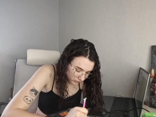 NatoshaBurdine, a white woman with curly black hair and glasses, leans over a laptop, writing with a pink pen. She has tattoos on her left arm and wears a black tank top. Fucking focused.