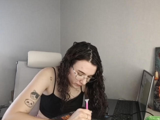 Natosha Burdine, eine blasse Frau mit langen, lockigen schwarzen Haaren, die eine Brille trägt und ein schwarzes BH trägt, sitzt an einem Schreibtisch. Laptop und Stift, mit Fokus auf das Schreiben. Tätowierung an ihrem Arm.