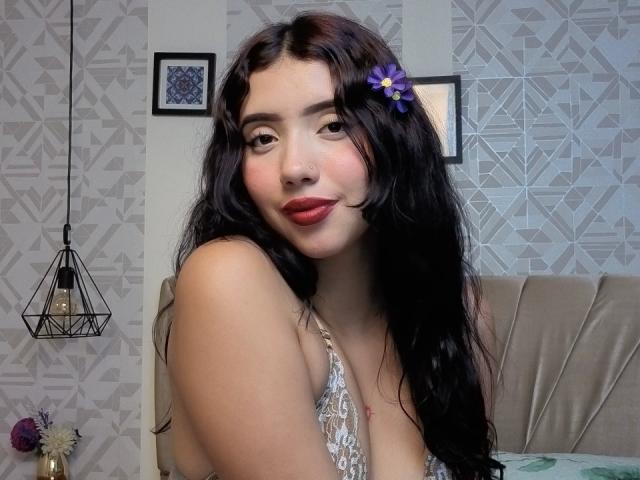 AbrilKoss with long black hair, wearing a silver lace top, has a blue flower in her hair, and a nose ring. She's smiling seductively, showing red lips. Geometric wallpaper background.