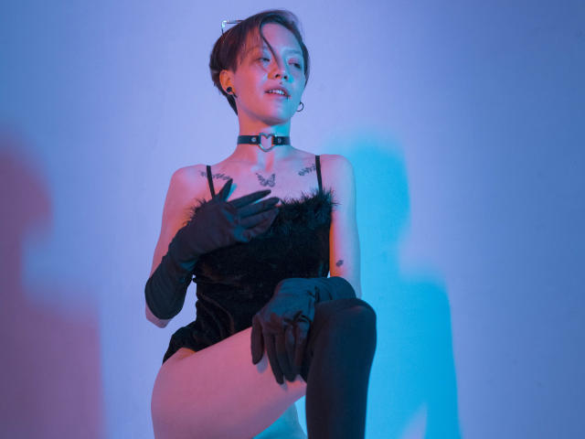 LissyLuxe adopte une pose séduisante, vêtue d'une robe noire ornée de plumes, de gants noirs et d'un collier. Des lumières bleues et violettes mettent en valeur sa poitrine et sa cuisse tatouées. Sa main repose sur sa cuisse.