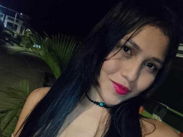 Nahaufnahme von Valentina Moreau, einer dunkelhaarigen Frau mit leuchtend rotem Lippenstift, einem blauen Choker, leicht geöffneten Lippen, verführerisch. Blicken. Dunkler Hintergrund, eine grüne Pflanze ist teilweise sichtbar. Schwach beleuchtet, intim, sexuelle Spannung..