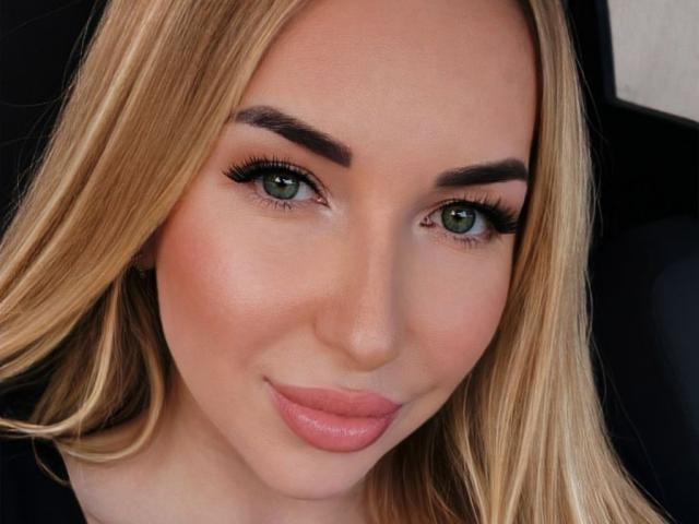Nahaufnahme von AriaGorgeous, einer blonden Frau mit grünen Augen, vollen, rosafarbenen Lippen und dicken, schwarzen Augenbrauen. Sie hat einen verführerischen Blick, betonten Eyeliner und ein leichtes Lächeln. Verdammt heiß.