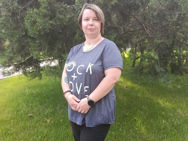 Photograph of BonnnyBoobsy, standing in a grassy area with trees. She has short brown hair, wearing a gray t-shirt with "COCK DIVE" in white letters, black pants, and a black watch. Her hands are clasped in front.