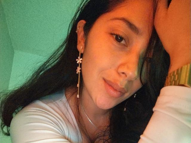 Primer plano de ChantalBernadette, una mujer con cabello largo y negro, que lleva una blusa blanca y aretes dorados, inclinando la cabeza. a la derecha, creando una expresión sugestiva y sensual. Fondo de color verde brillante.