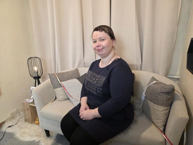 BonnnyBoobsy sits on a light gray couch, wearing a black long-sleeve top and black pants. She's smiling, with hands resting on her lap. Soft lighting, white curtains in background.