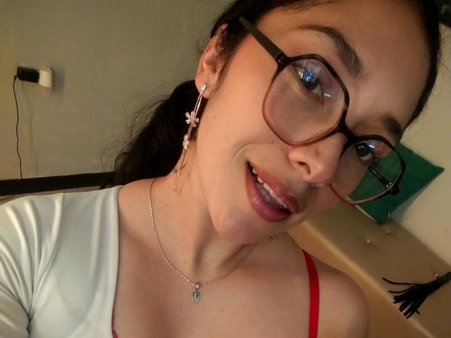 Close-up of ChantalBernadette with black hair, glasses, and red strap visible on white top, smiling with slightly open mouth. She's wearing a necklace and star-shaped earrings, looking directly at camera.