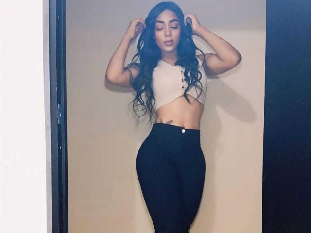 PaisitaBlack è in piedi di fronte a una parete beige, con le mani tra i lunghi capelli neri, indossa un top bianco corto e pantaloni neri a vita alta. pantaloni, occhi chiusi, postura sicura, figura snella, sexy, sicuro di sé.