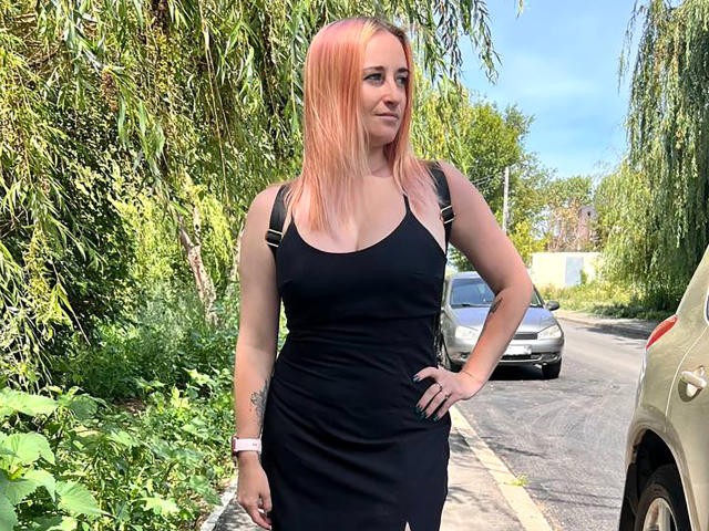 MeiraGilson, una mujer rubia con cabello lacio, vestida con un vestido negro ajustado, de pie en una calle soleada, con una mano en la cadera, coche. y exuberante vegetación en el fondo, pose casual y sensual..