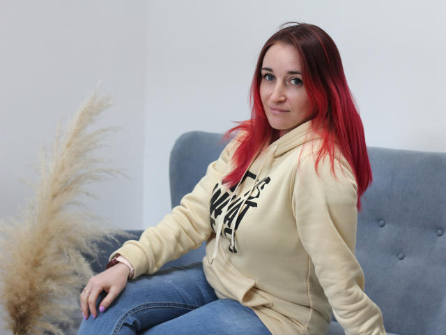 MeiraGilson, with red hair, sits on a blue couch, wearing a yellow hoodie and blue jeans. Her hand rests on a puffed-up cushion, creating a relaxed, slightly provocative pose.