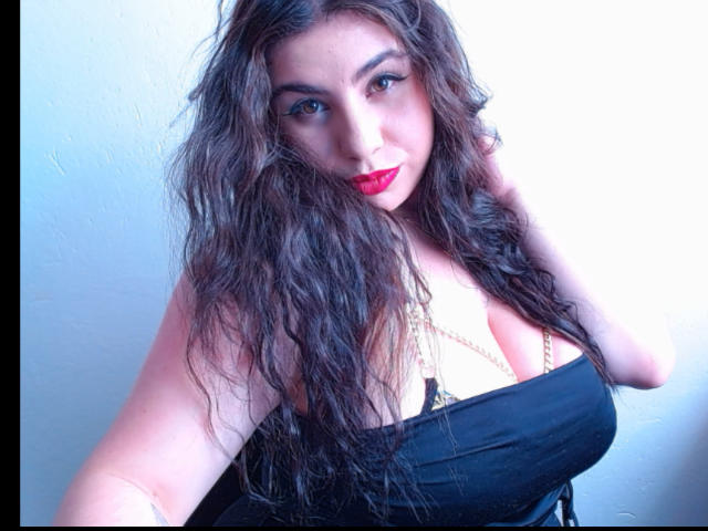 Fotografia de VenusxMusa, uma mulher curvilínea com longos cabelos castanhos ondulados, vestindo um top preto sem alças, que expõe o colo. fenda. Olhos azuis brilhantes, batom vermelho, cabelo levemente despenteado, olhar intenso, iluminação forte..