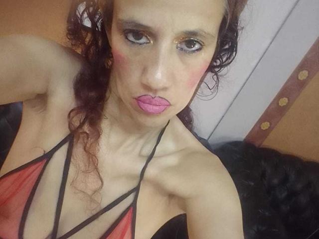 Gros plan de NatashaDirk, à la peau pâle et aux cheveux bruns bouclés, portant un haut rouge à encolure américaine, du rouge à lèvres rose et un maquillage des yeux "smoky". levant les yeux avec une expression légèrement séduisante, une bretelle noire visible..