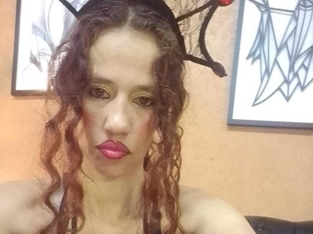 Fotografia di Natasha Dirk con lunghi capelli castani ricci, che indossa fasce per capelli nere a forma di corna e un rossetto rosa acceso, mentre guarda. Fissando direttamente la telecamera con un'espressione seria. Incredibilmente attraente, con un intenso contatto visivo..