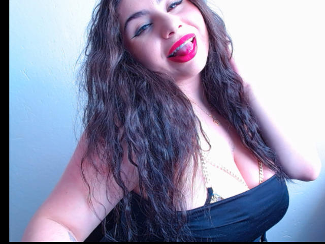 Close-up of VenusxMusa with long, wavy brown hair, red lipstick, and black strapless top. Bright light highlights her cleavage. She's tilting head, looking up with a seductive smile. Overexposed background.