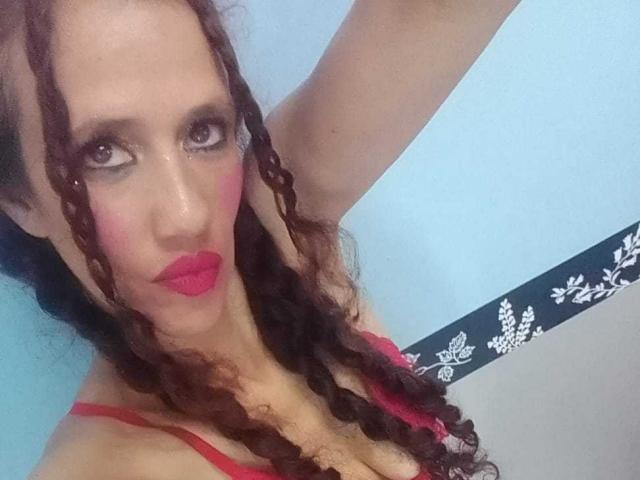 Nahaufnahme von NatashaDirk mit langen, dunklen Zöpfen, rotem Lippenstift und einem roten BH. Sie blickt verführerisch, ihre Hand ist erhoben. Ihre Brüste sind deutlich sichtbar und erzeugen eine sexy, provokante Atmosphäre..