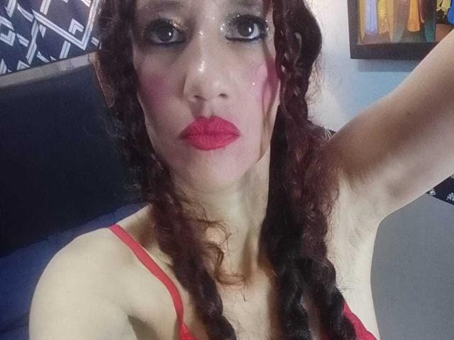 Close-up selfie of NatashaDirk, face covered in a shiny, translucent substance. She has curly brown hair, red lipstick, and wears a red top. Arm raised, intense gaze, sexual tension.