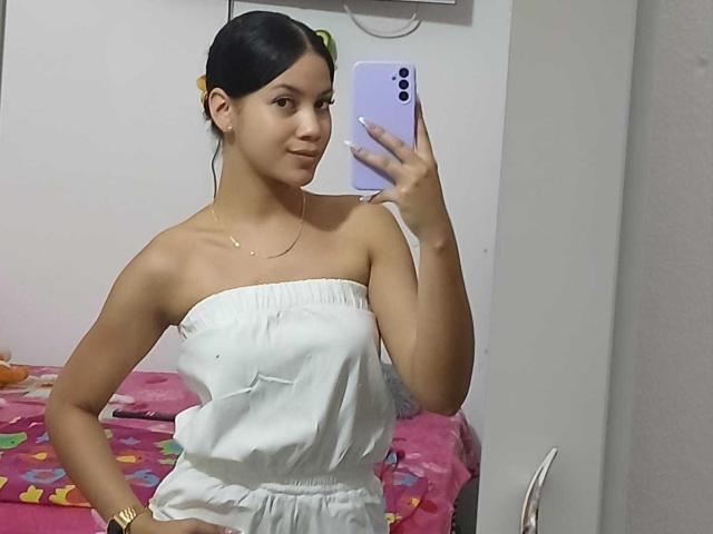 AliceFoxxy si fa un selfie con un iPhone bianco, indossando una canottiera bianca senza spalline, mostrando le sue spalle scoperte e un accenno di scollatura. I suoi capelli scuri sono legati indietro, e si trova in una camera da letto con una coperta rosa..